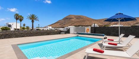 Une piscine entourée de chaises longues et d’un parasol, le tout bordé d’un mur de pierre et de palmiers, avec une montagne en arrière-plan.