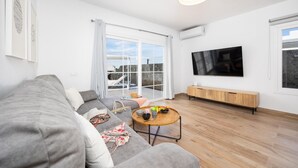 A modern living room with a grey sofa, a coffee table with fruits, a TV, and a large window with a view of buildings. - Villa Yaiza Secret 2 (Yaiza)