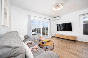 Ein modernes Wohnzimmer mit einem grauen Sofa, einem Couchtisch mit Früchten, einem Fernseher und einem großen Fenster mit Blick auf Gebäude.