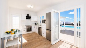 A modern kitchen with white cabinets, a dining table with chairs, and a view of a swimming pool through an open door. - Villa Yaiza Secret 2 (Yaiza)