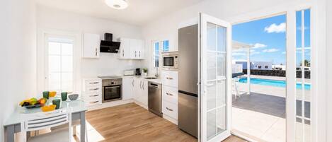 A modern kitchen with white cabinets, a dining table with chairs, and a view of a swimming pool through an open door.