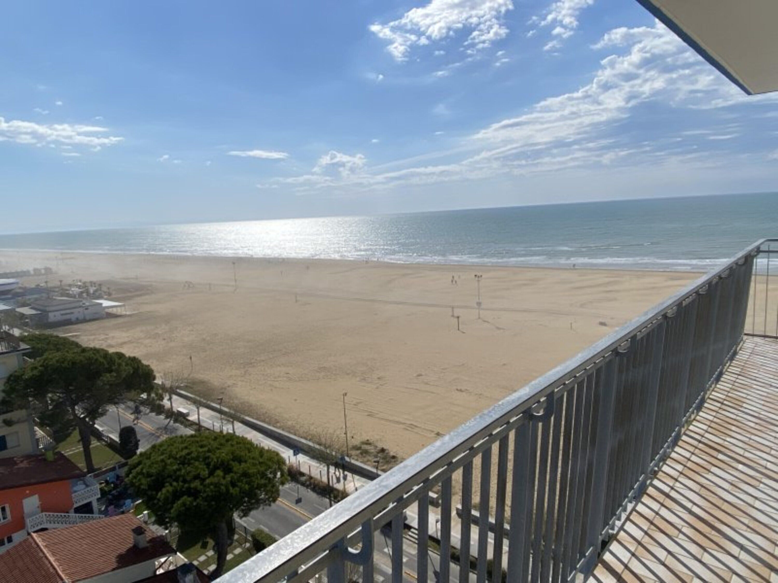 A beach view from a balcony with a railing, a road with vehicles, and buildings in the foreground.