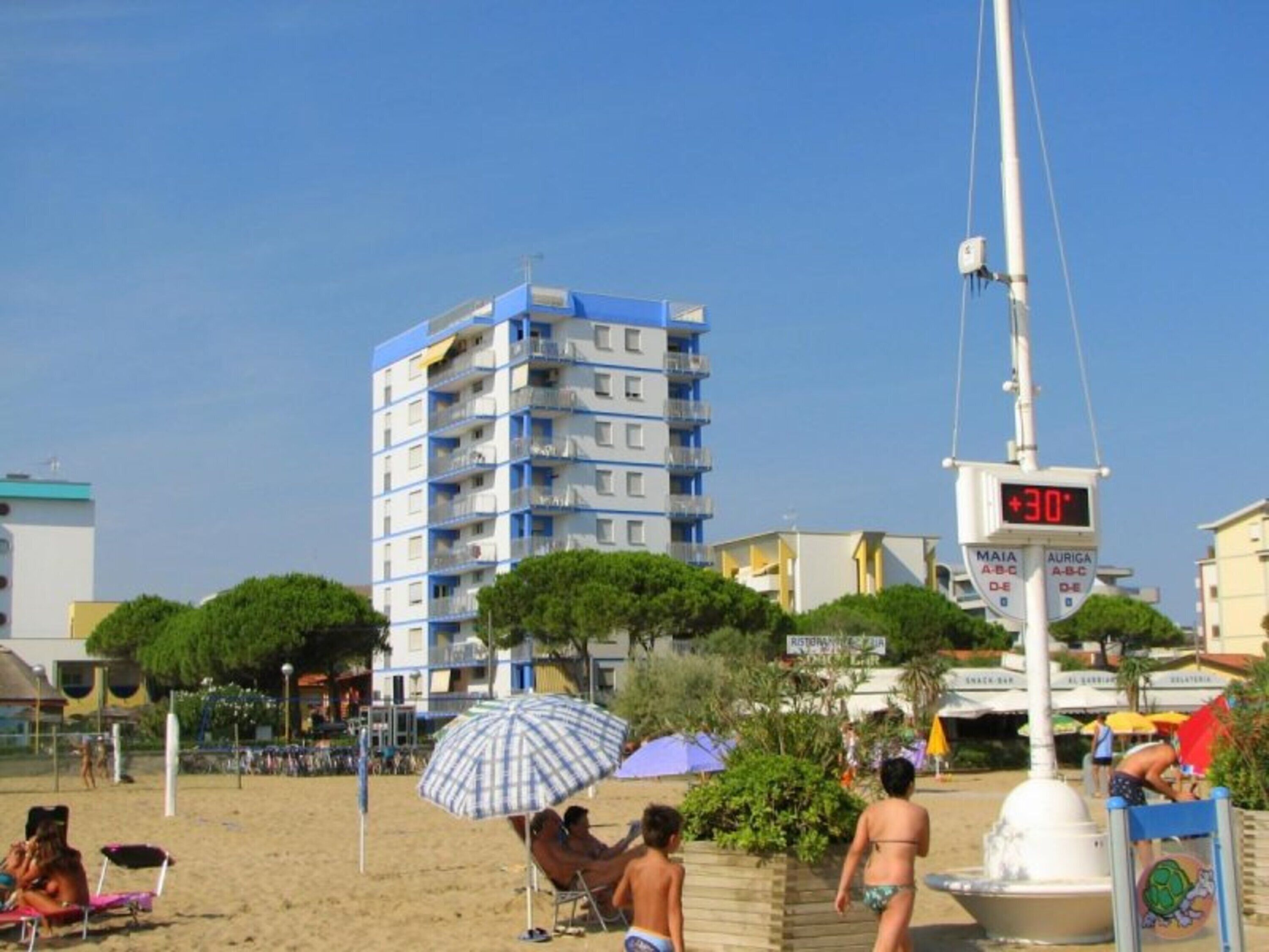 Una playa con un edificio alto, un indicador digital de temperatura y un cartel que muestra 'MAIA' y 'AURIGA', indicando lugares cercanos.