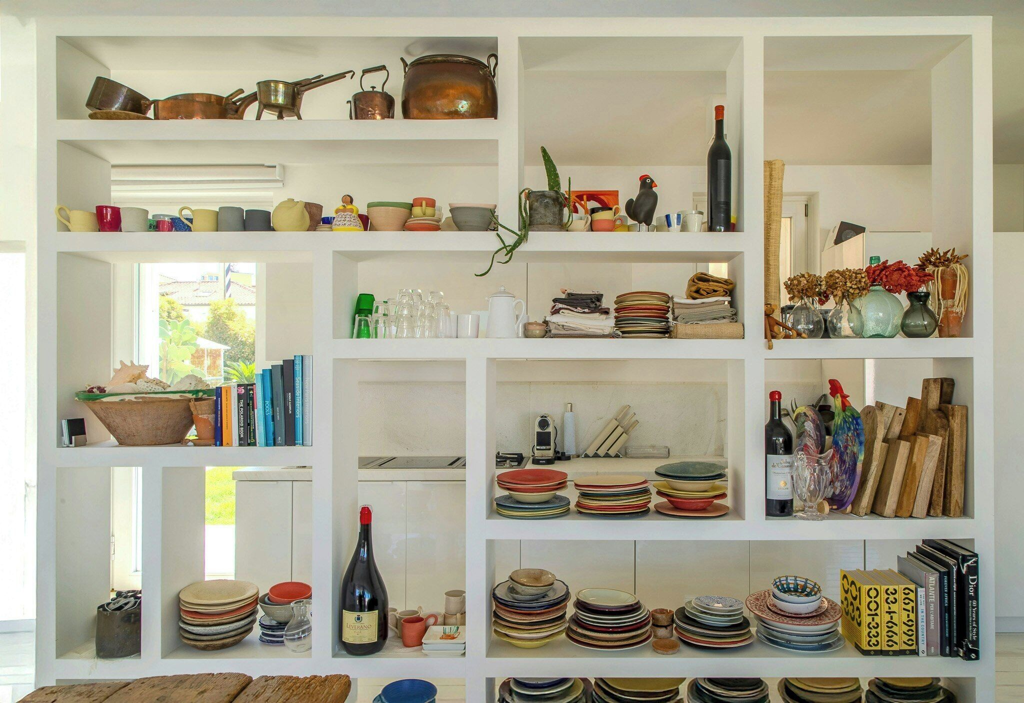 A kitchen with shelves containing various kitchenware, a wine bottle, and a window with a view of greenery.