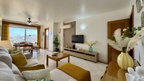A living room with a sofa, coffee table, TV, and a dining area with a table and chairs. - Albufeira Amazing View 2 by Homing (Albufeira)