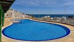 A swimming pool with a clear view of the sea, a covered area with lounge chairs, and a building with a white facade. - Albufeira Amazing View 2 by Homing (Albufeira)