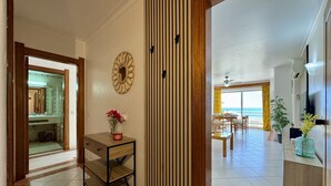 A hallway leading to a dining area with a view of the ocean, a wooden cabinet with a vase of flowers, and a mirror on the wall. - Albufeira Amazing View 2 by Homing (Albufeira)