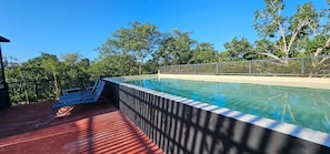 Una piscina con una valla metálica, un banco y árboles al fondo.