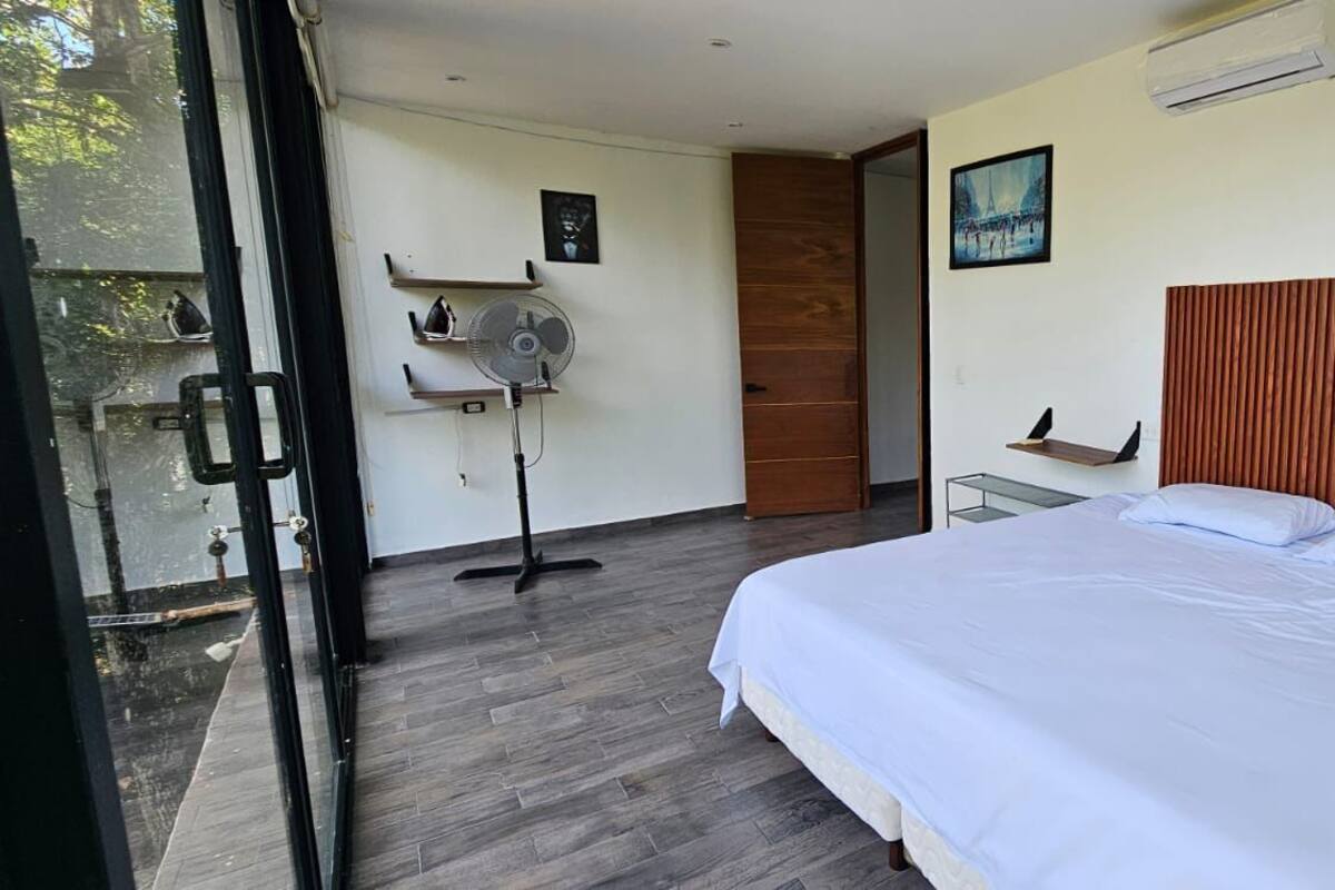 A bedroom with a bed, a standing fan, a wooden door, and a window.