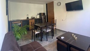 Una sala de estar con un sofá marrón, una mesa de comedor con sillas, una zona de cocina con un refrigerador y un televisor de pantalla plana montado en la pared.