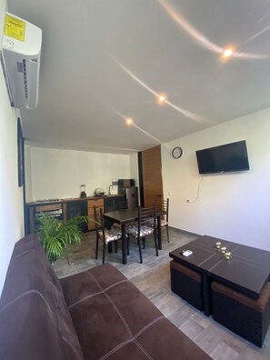 A living room with a brown sofa, a dining table with chairs, a kitchen area with a refrigerator, and a flat-screen TV mounted on the wall. - Cuatro Elementos Noh By GEstores (Tulum)