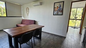 Una habitación con una mesa de madera, dos sillas, un banco acolchado rojo, un aire acondicionado de pared, un cuadro enmarcado y un ventanal grande.
