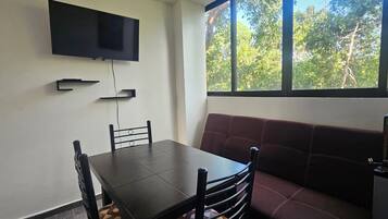 Una habitación con una televisión montada en la pared, una mesa con sillas, un sofá y un ventanal con vistas a árboles.