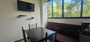 Una habitación con una televisión montada en la pared, una mesa con sillas, un sofá y un ventanal con vistas a árboles.