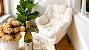 Una habitación acogedora con un sillón blanco, un cuadro de una pareja en la playa, una botella de vino y un jarrón de flores.