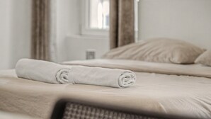 Cama bem arrumada com toalha enrolada, janela com cortinas e parede lisa.