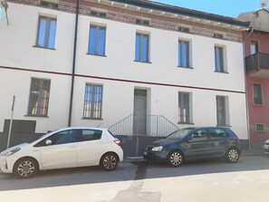 A two-story building with a gated entrance, a white car parked in front, and a dark-colored car parked beside it. - Apartment 'Blu' with Wi-Fi and Air Conditioning (Delebio)