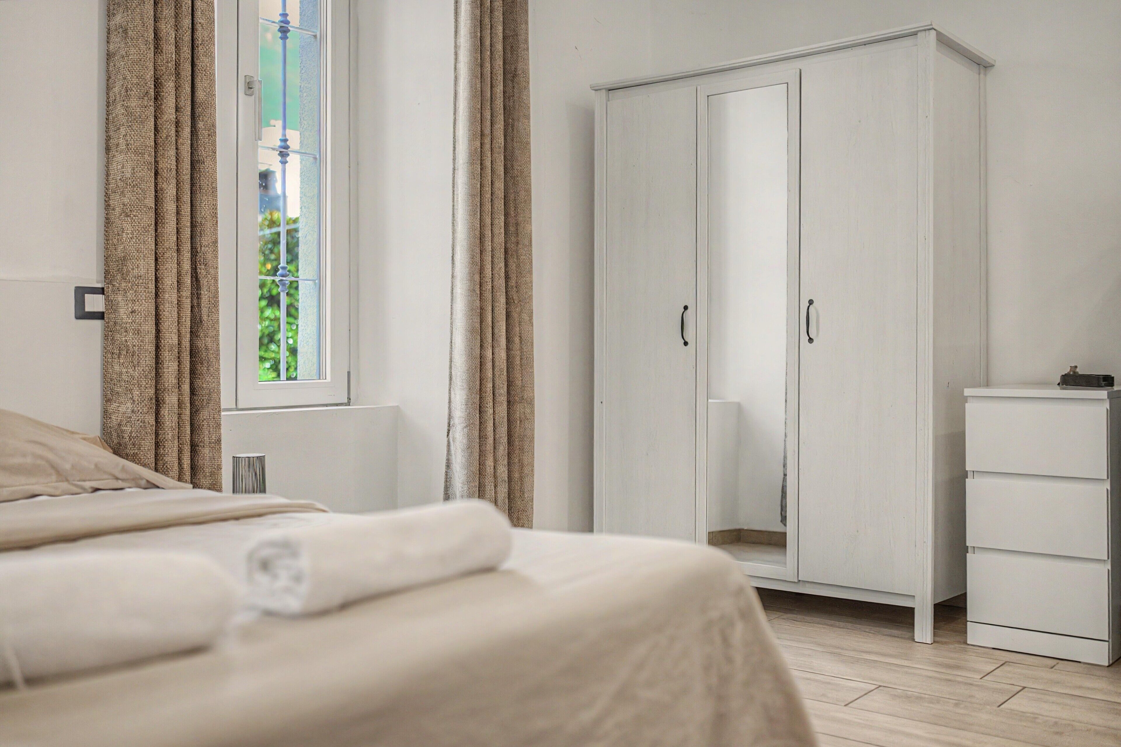 A bedroom with a bed, a wardrobe, a bedside table, and a window with curtains.