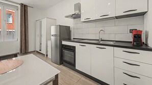 Uma cozinha moderna com armários brancos, uma geladeira de aço inoxidável, forno embutido, máquina de café e uma janela com cortinas.