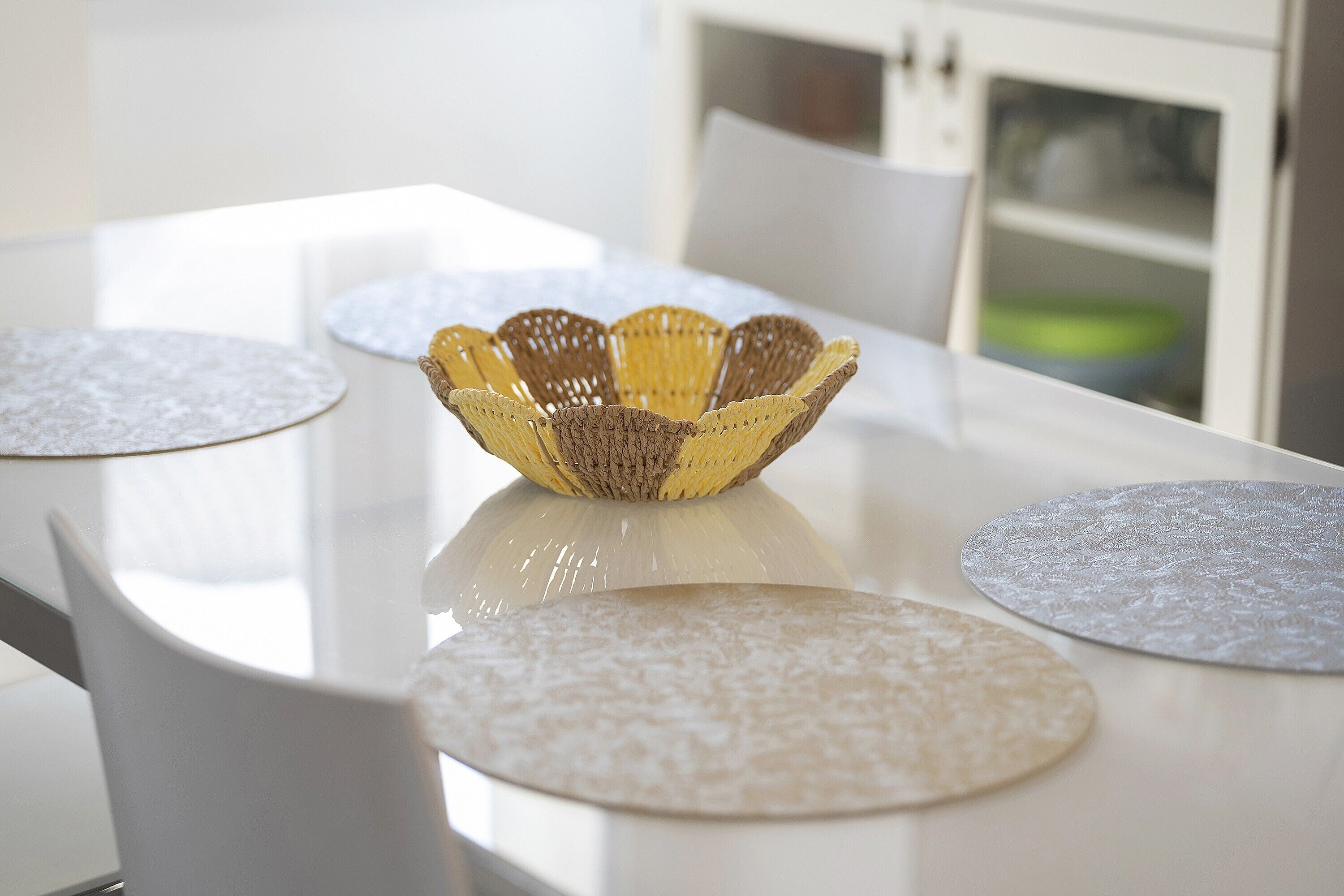 A dining table with a woven centerpiece, white chairs, and round placemats.