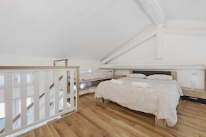 Une chambre avec un plancher en bois, un lit recouvert d’une courtepointe blanche, une table de chevet et une balustrade en bois.