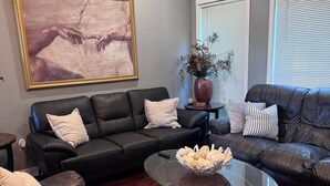 A living room with two black leather sofas, a glass coffee table, a painting on the wall, and a vase with dried flowers on a side table. - Creative Heart - Get Away!!   Spacious 4BR/3 Full Bath Home for your Enjoyment. (Gulfport)