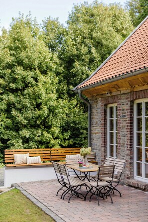 Eine Terrasse mit einem Ziegel-Tisch und -Stühlen, einer hölzernen Bank mit Kissen sowie einem Steinhaus mit roten Ziegeldach.