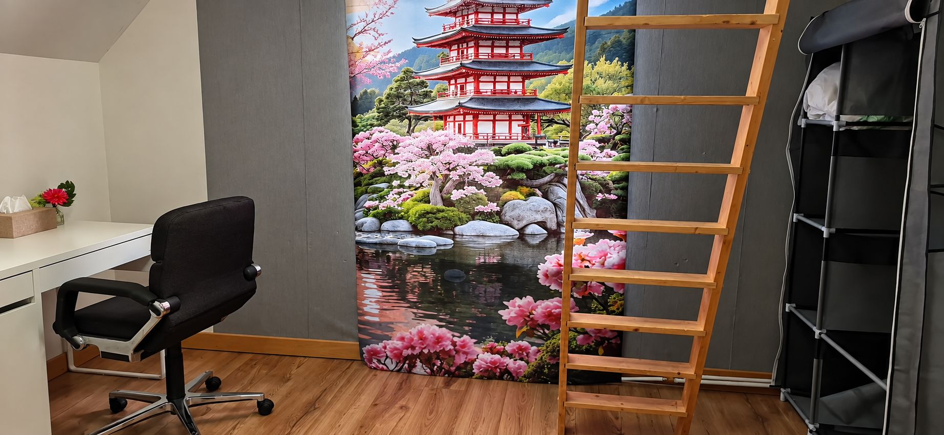 Una habitación con un escritorio, una silla de oficina negra, una escalera de madera y una pared con un mural panorámico de una pagoda y flores de cerezo.