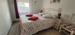 Ein Bett mit floraler Bettwäsche, zwei rote Kissen, ein Nachttisch mit Lampe und ein kleiner Stuhl mit gestreifter Sitzkissen.