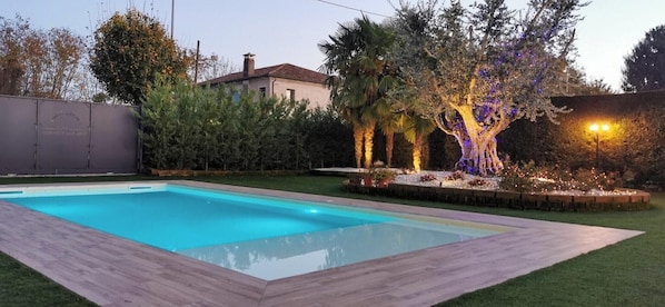 Une piscine rectangulaire dotée d’une terrasse en bois, entourée d’une pelouse bien entretenue et de différents types d’arbres.
