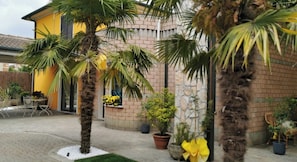Un bâtiment à la façade jaune, avec un mur en briques et un toit recouvert de tuiles. On y trouve des plantes en pot et un petit espace patio aménagé avec du mobilier d’extérieur.