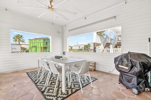A patio area with a table and chairs, a ceiling fan, a grill, and a view of neighboring buildings. - Resting Beach Face | Private Pool • Modern Coastal Retreat • Prime 11th St Location (Port Aransas)