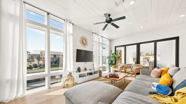 A spacious living room with a large gray sofa, a flat-screen TV, and a ceiling fan. - Resting Beach Face | Private Pool • Modern Coastal Retreat • Prime 11th St Location (Port Aransas)