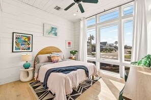 A bedroom with a bed, a nightstand, a lamp, a ceiling fan, and a large window with a view of the outside. - Resting Beach Face | Private Pool • Modern Coastal Retreat • Prime 11th St Location (Port Aransas)
