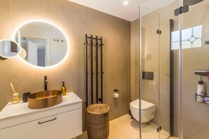 Un baño moderno con un espejo redondo, un lavabo con pileta, un inodoro y una ducha con mampara de vidrio.