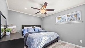 Ein Schlafzimmer mit Bett, Deckenventilator, Fenster mit Jalousien, Spiegel und Kommode.