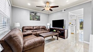 Ein Wohnzimmer mit braunem Ledersofa, einem Couchtisch, einem Flachbildfernseher, einem Deckenventilator und einem Gemälde an der Wand.