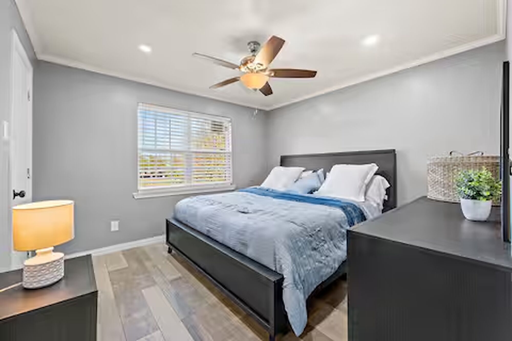 Ein Schlafzimmer mit einem Bett, einem Deckenventilator, einem Fenster mit Jalousien, einem Nachttisch mit Lampe und einer Kommode.