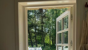 Uma porta aberta que dá para um deck de madeira com guarda-corpo branco, uma toalha pendurada e vista para uma área arborizada.