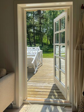 Eine offene Tür, die auf eine Holzterrasse mit weißer Brüstung hinausführt, wo ein Handtuch hängt und der Blick auf eine bewaldete Gegend fällt.