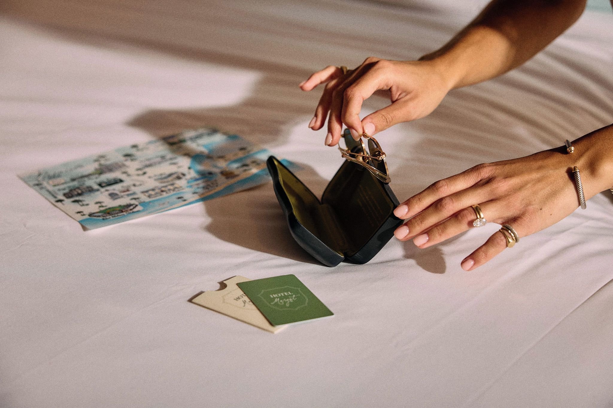 Uma pessoa segurando uma carteira preta com fecho dourado, deitada em uma cama com uma revista e um folheto de hotel.