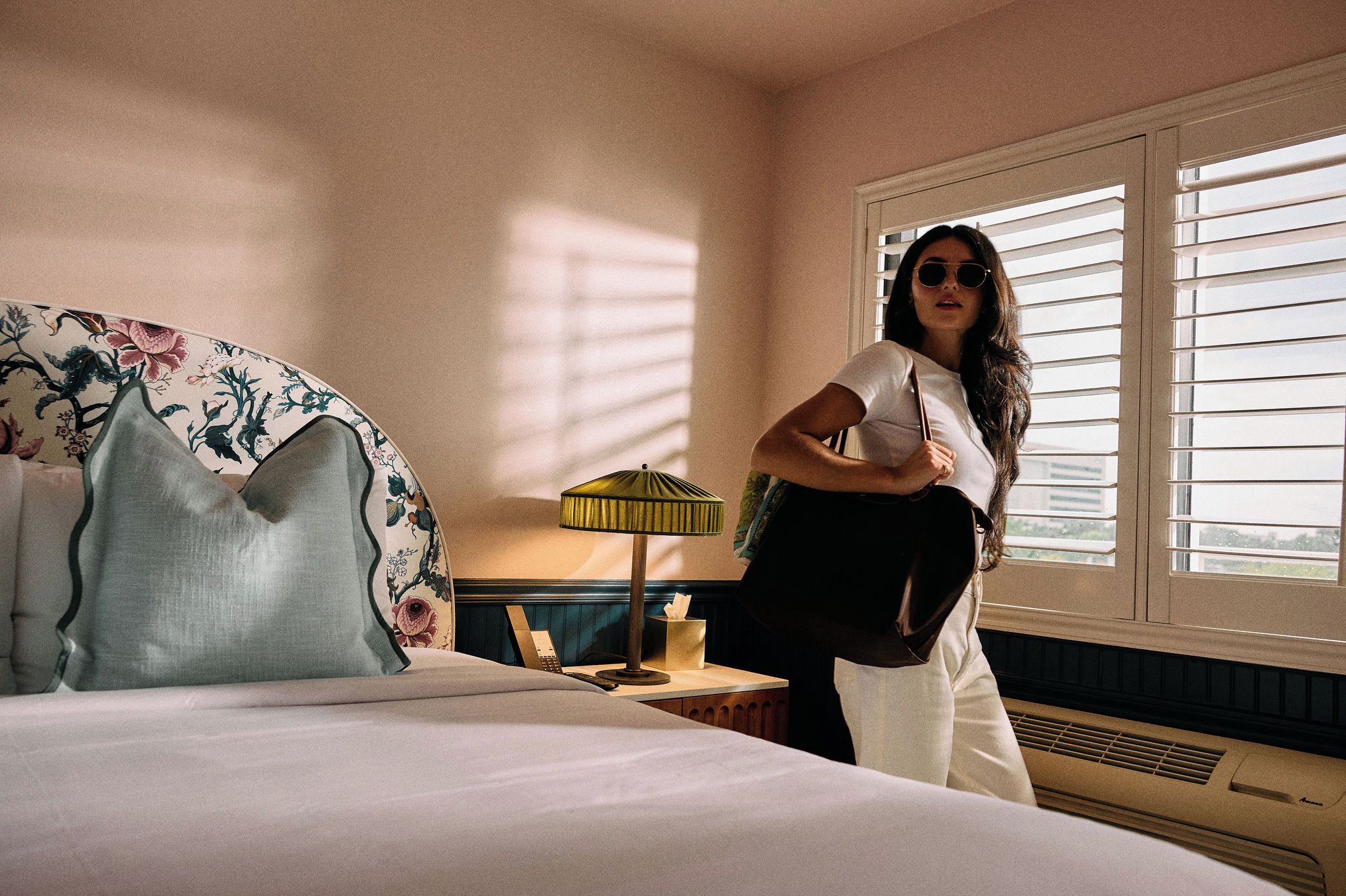 A hotel room with a bed, a nightstand, a lamp, and a window with blinds.