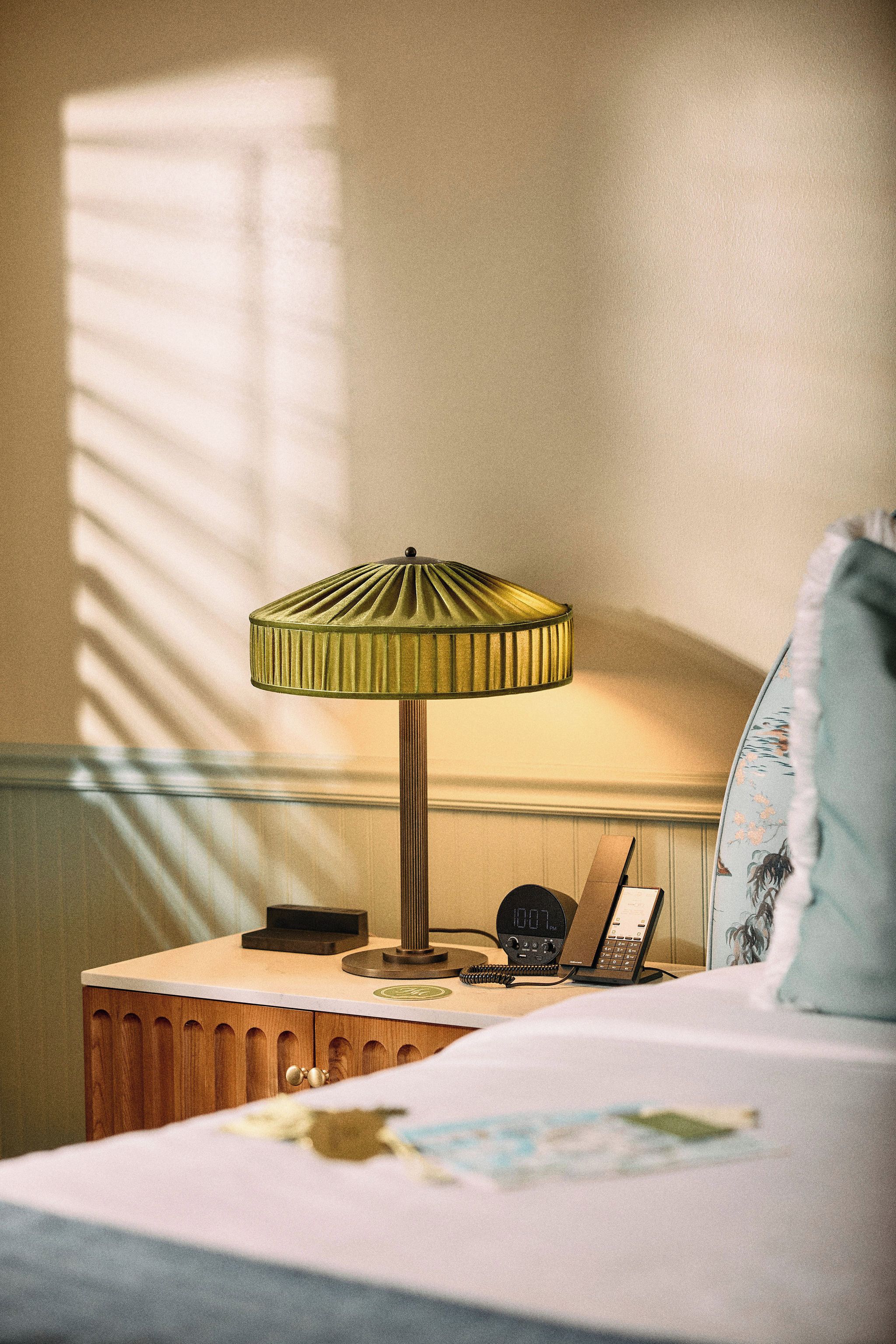 Una habitación de hotel con una cama, una mesita de noche con lámpara, un teléfono y un reloj.