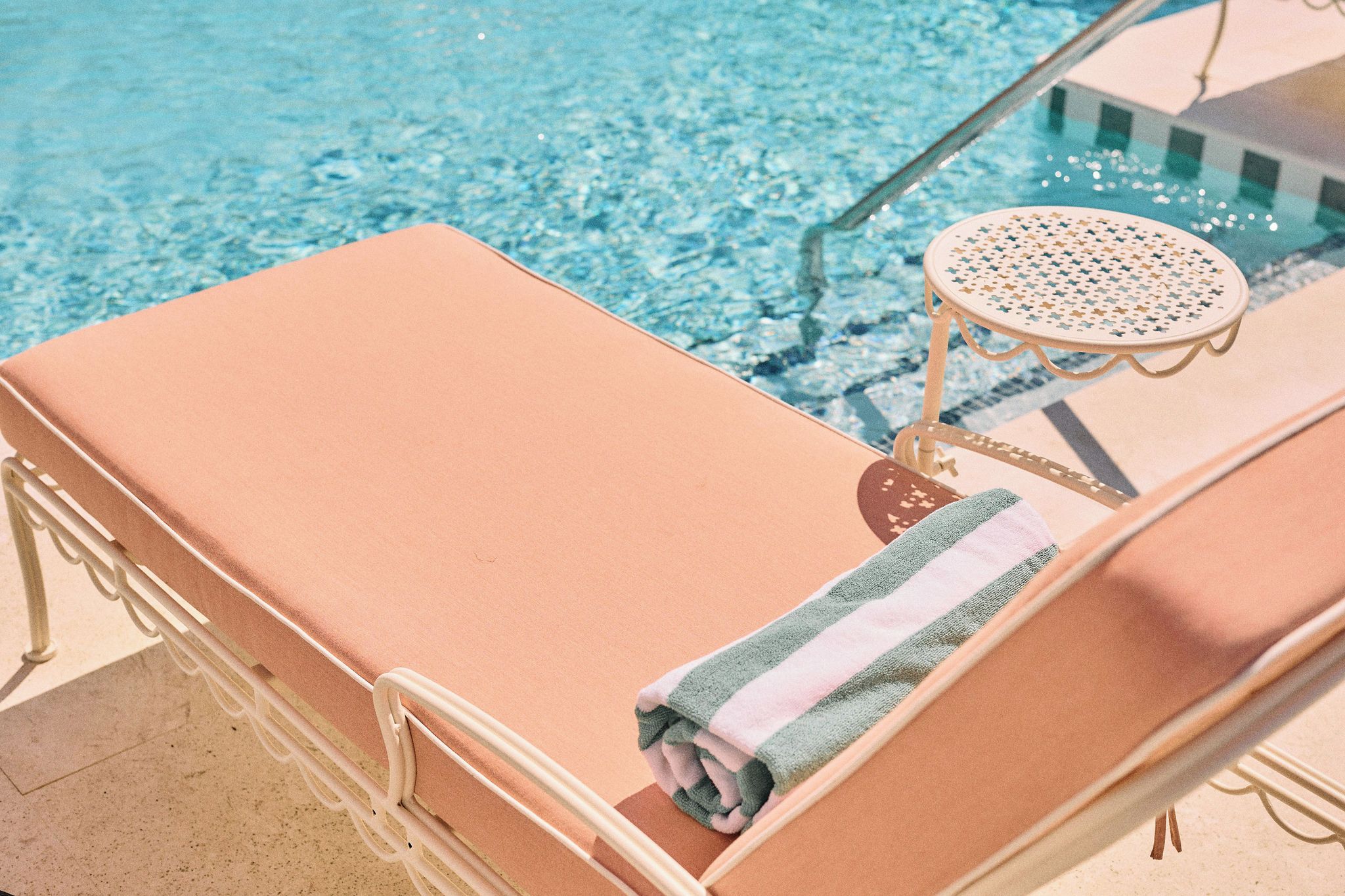 Una cadira de relax a la piscina amb una tovallola, una taula rodona petita amb un disseny perforat, i una piscina amb aigua blava i cristal·lina.