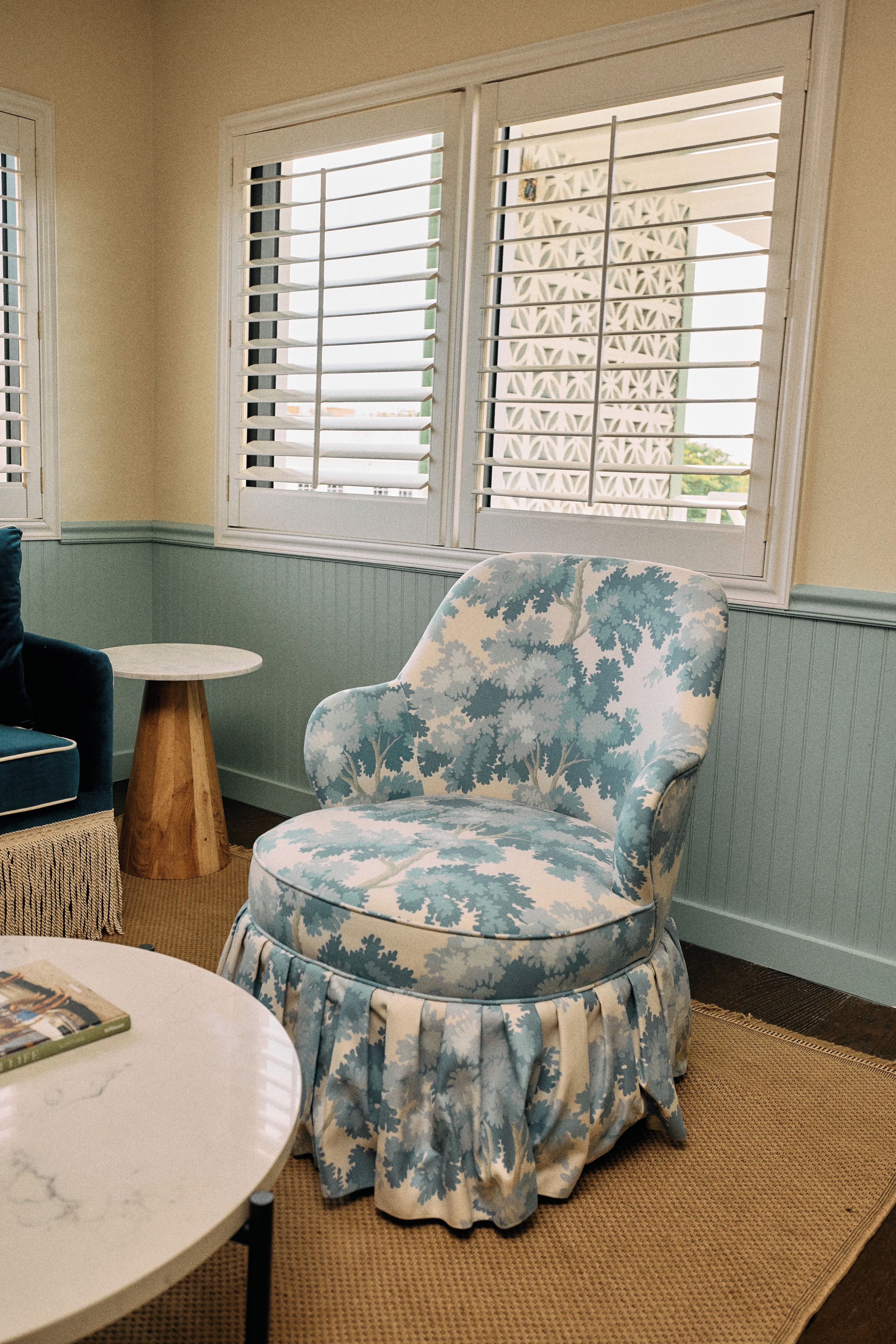 A room with a patterned armchair, a round table, and a window with blinds.