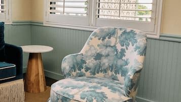 A room with a patterned armchair, a round table, and a window with blinds.