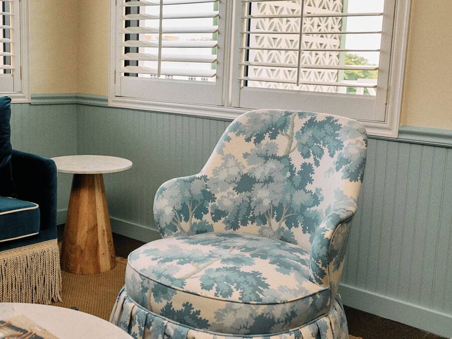 A room with a patterned armchair, a round table, and a window with blinds.
