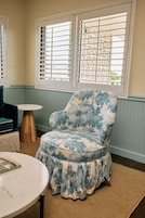 A room with a patterned armchair, a round table, and a window with blinds.