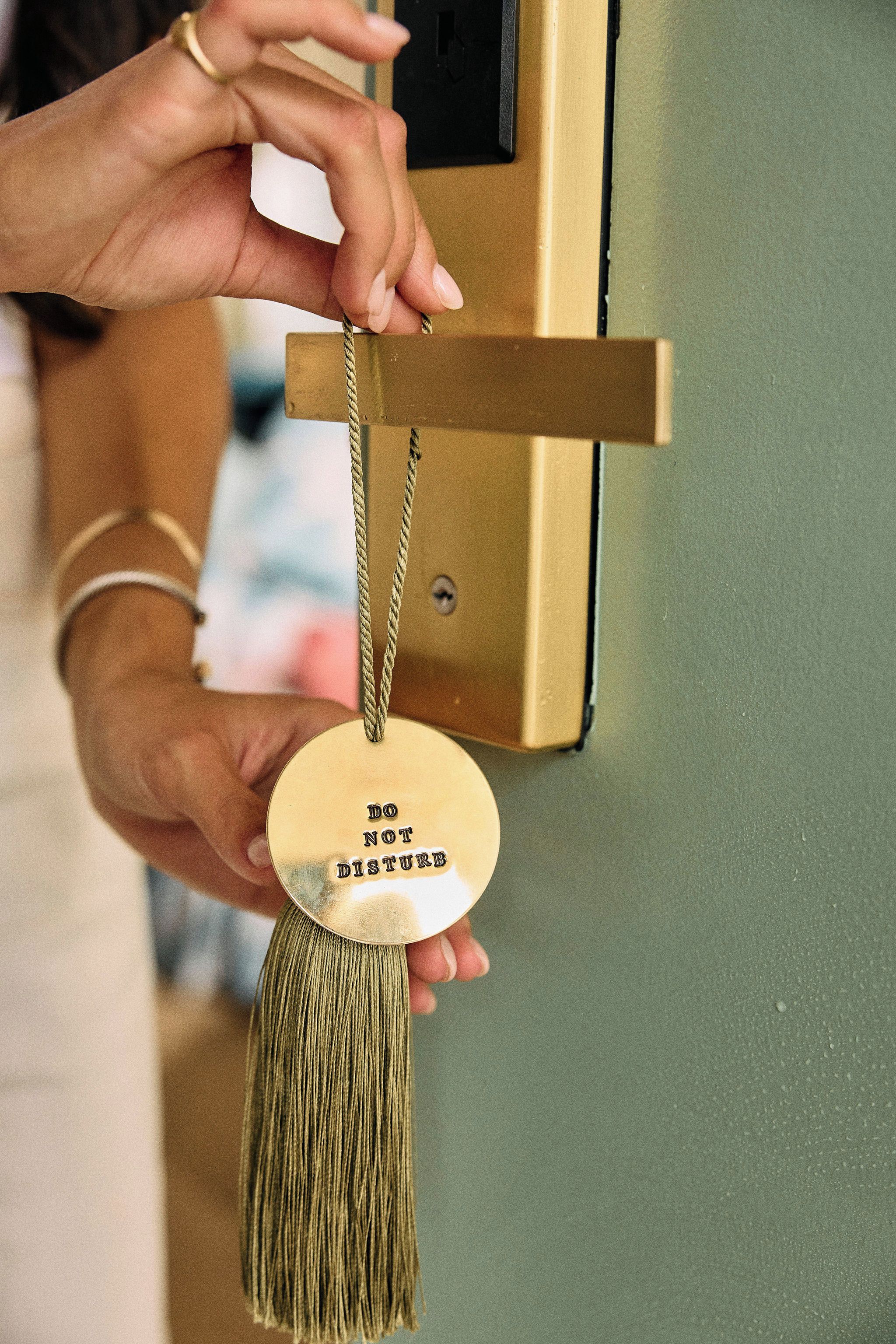 A hand holding a round, gold-colored keychain with a tassel and the inscription 'DO NOT DISTURB' on a door lock.