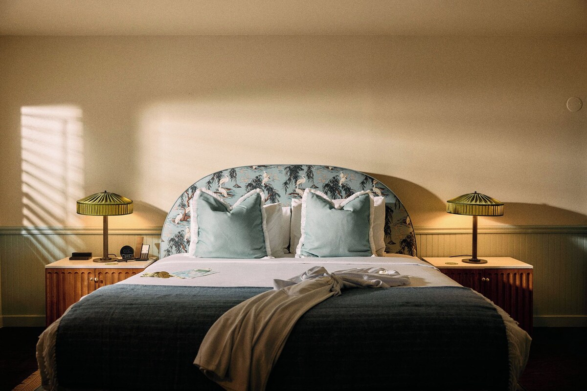 Una cama bien hecha con cabecera floral, dos lámparas de noche y una mesita de noche de madera con un reloj y un teléfono.